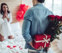 Beautiful young couple in bedroom. Celebrating Saint Valentine's Day with gift box, air baloons in shape of heart and red roses.